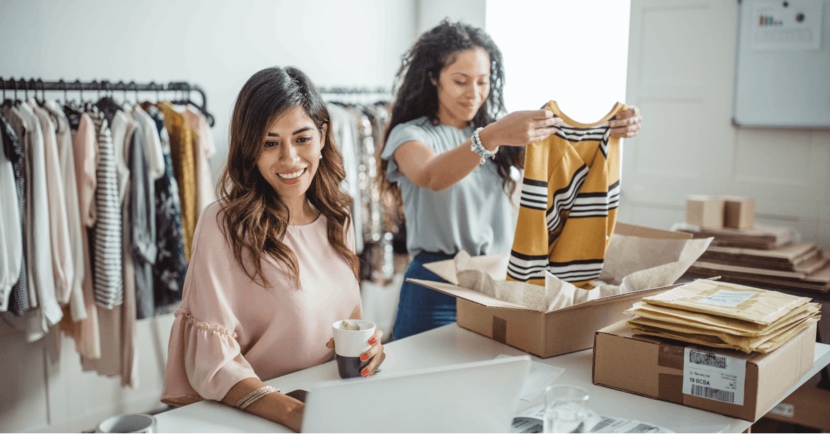 two women working at table packing clothes into cardboard boxes cup of tea laptop in foreground clothing racks in blurred background